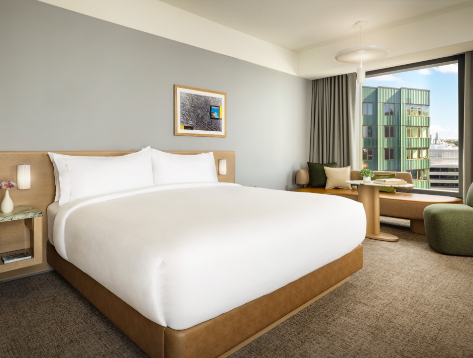 King-size bed with white bedding and a light wood headboard, a small bedside table with a vase of flowers, and a framed picture on a gray wall in a bright hotel room.