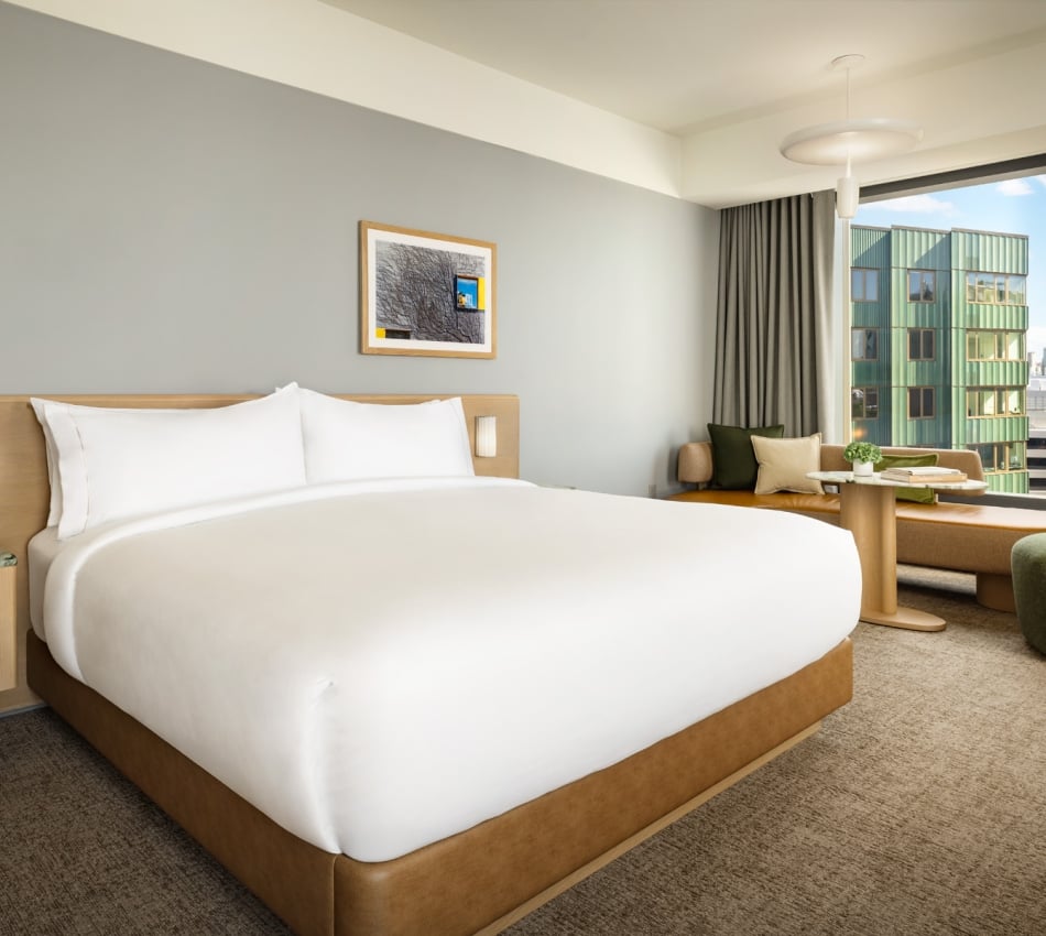 King-size bed with white bedding and a light wood headboard, a small bedside table with a vase of flowers, and a framed picture on a gray wall in a bright hotel room.