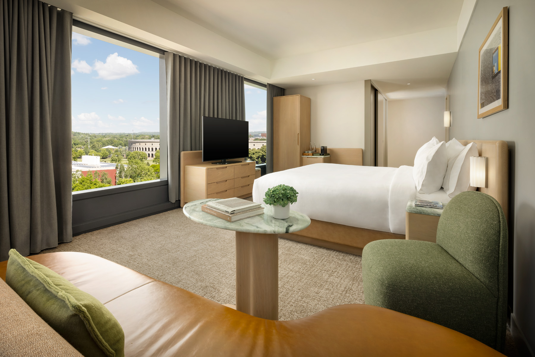 Spacious hotel suite living area with a white bed, green chairs, and a round marble table overlooking a city view through large windows.
