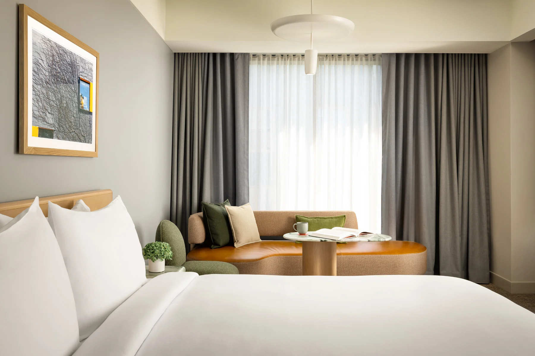 Hotel room living area: bed in foreground, beige sofa with green and beige cushions, round coffee table, and a large window with sheer and dark curtains.