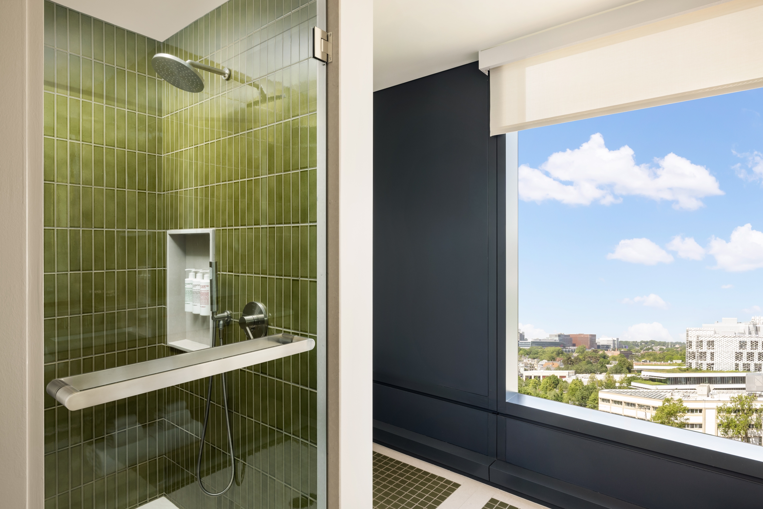 Shower stall with olive-green tile walls and chrome fixtures, opening to a city skyline on a sunny day.