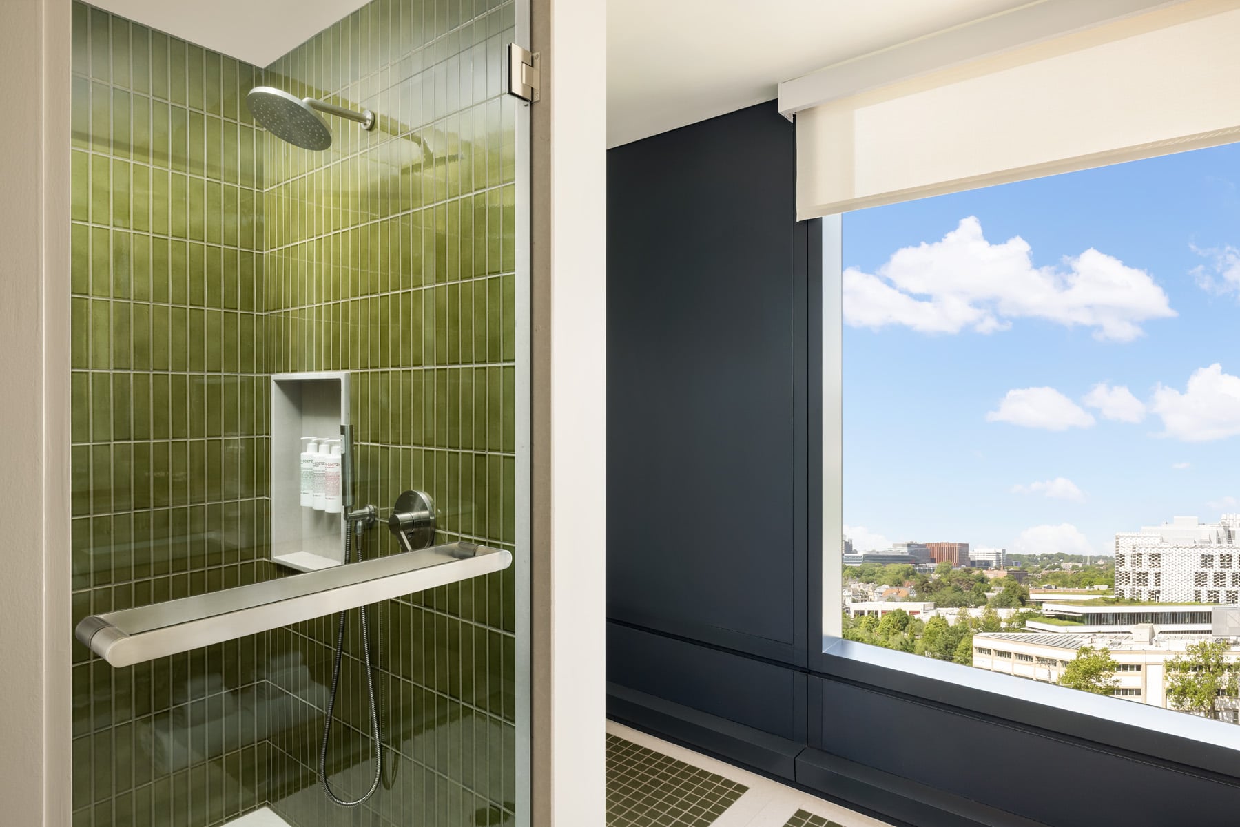 Shower area with green tiled walls, handheld shower hose, and a recessed shampoo niche, overlooking a cityscape through a large window.