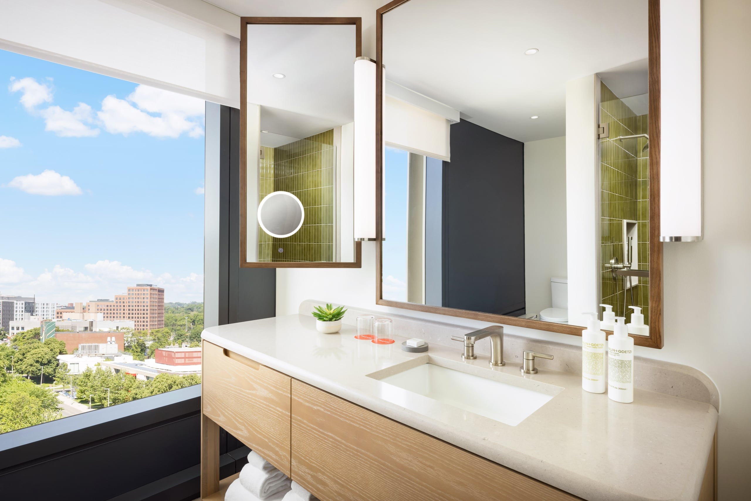 Modern bathroom with large windows, wood-framed mirrors, a green-tiled shower, and a white countertop with lotion bottles on the sink area.