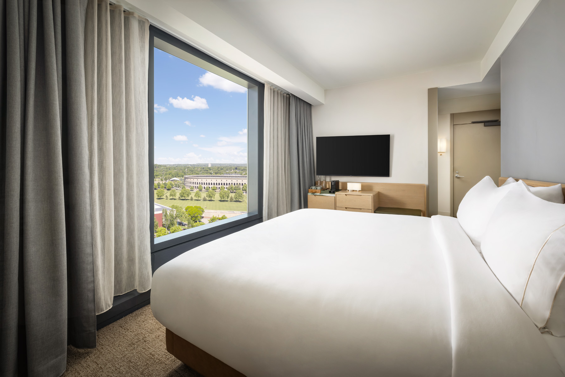 Hotel room with white bed, big window and gray curtains, city and park view outside.