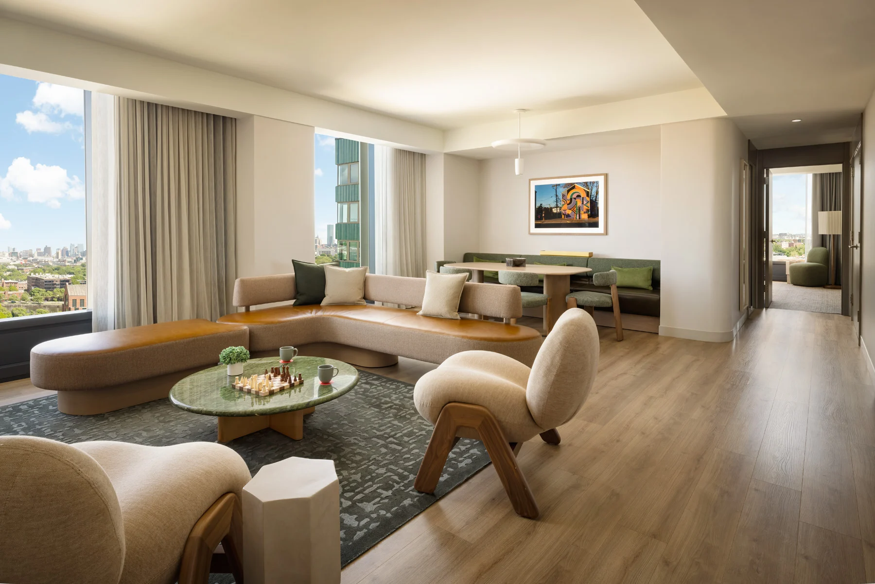 Bright open living area with a curved tan sofa, round glass coffee table, and large windows showing a cityscape.