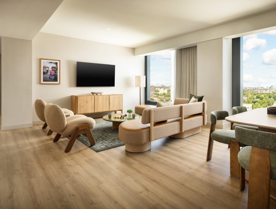 Bright, modern living room with beige sofa and chairs, a wall-mounted TV, and a city view through large windows.