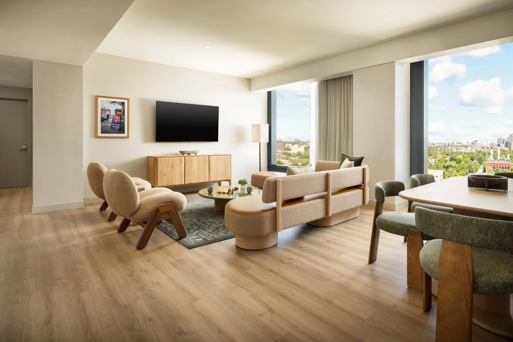 Bright, modern living room with beige sofa and chairs, a wall-mounted TV, and a city view through large windows.