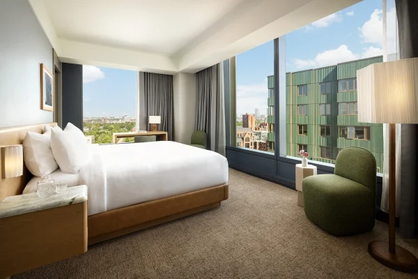 Sunlit hotel room with a king bed, white linens, wooden headboard, and city view through large windows.
