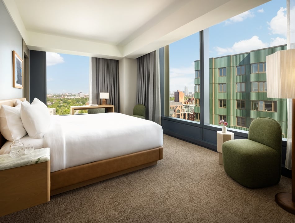Sunlit hotel room with a king bed, white linens, wooden headboard, and city view through large windows.