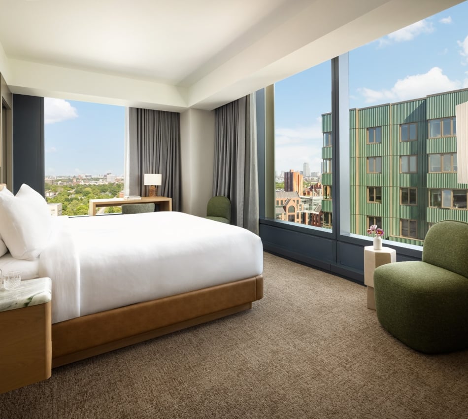 Sunlit hotel room with a king bed, white linens, wooden headboard, and city view through large windows.