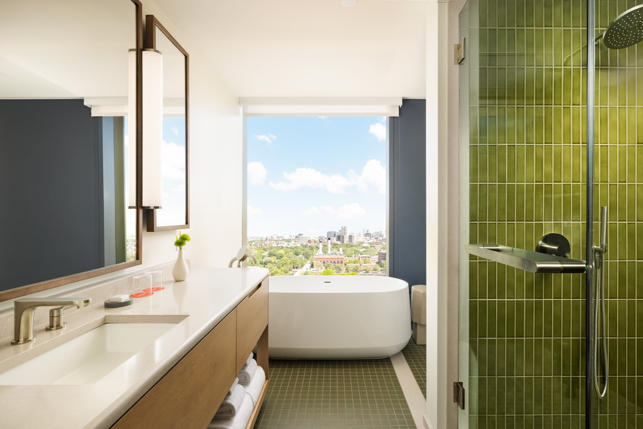 Bright bathroom with white countertop, wooden vanity, and freestanding tub facing a city view through a large window.