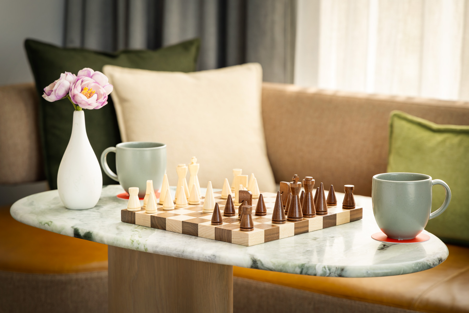 Vase with pink flowers, a chessboard with pieces, and two mugs on a round marble coffee table in a cozy living room setting.