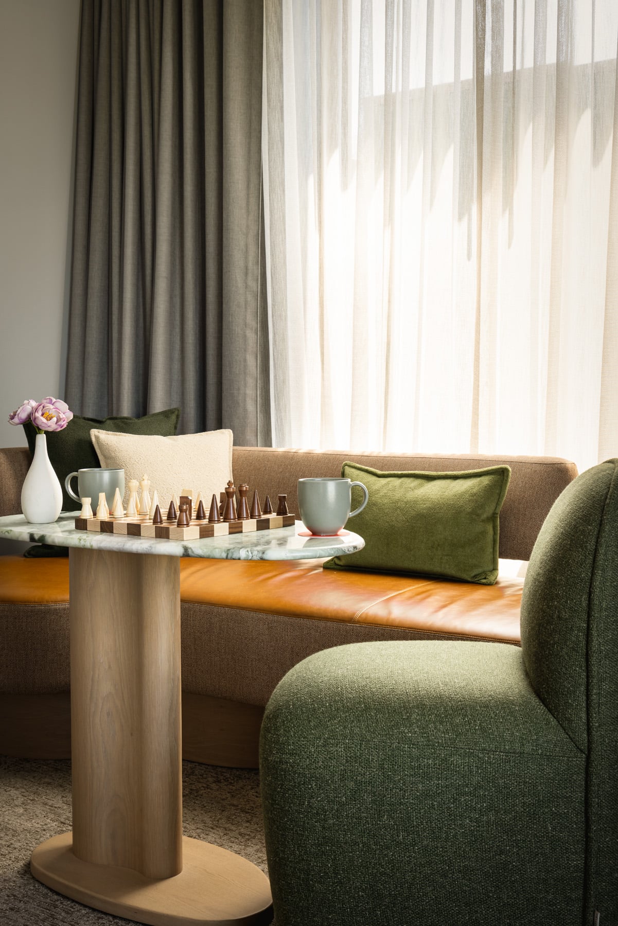 Cozy seating area with a marble round table, a wooden chessboard, and green cushions on a brown sofa by sheer curtains and a vase of pink flowers.