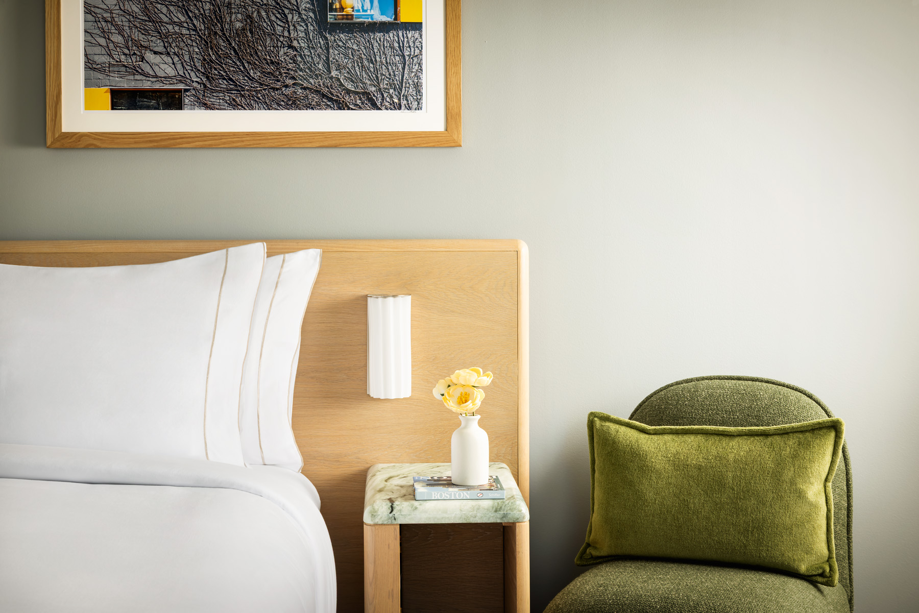 Bedroom scene with a white bed, wooden headboard, and a nightstand holding a vase of yellow flowers; a green upholstered chair to the right.