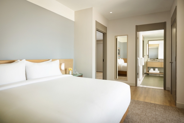 Hotel bedroom with a large white bed, wooden headboard, and a side table with a small vase of yellow flowers; doorway and mirrors on the wall to the right.