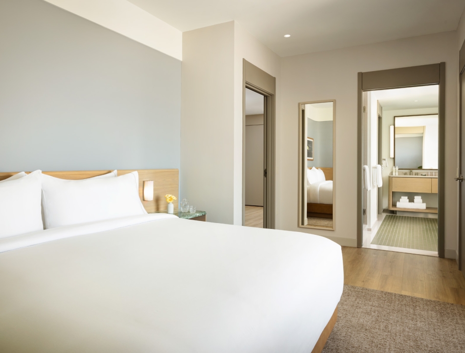 Hotel bedroom with a large white bed, wooden headboard, and a side table with a small vase of yellow flowers; doorway and mirrors on the wall to the right.