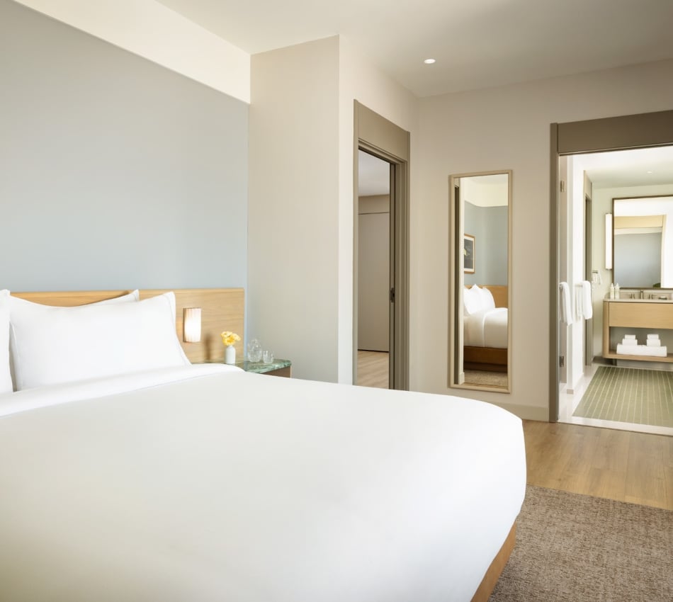 Hotel bedroom with a large white bed, wooden headboard, and a side table with a small vase of yellow flowers; doorway and mirrors on the wall to the right.