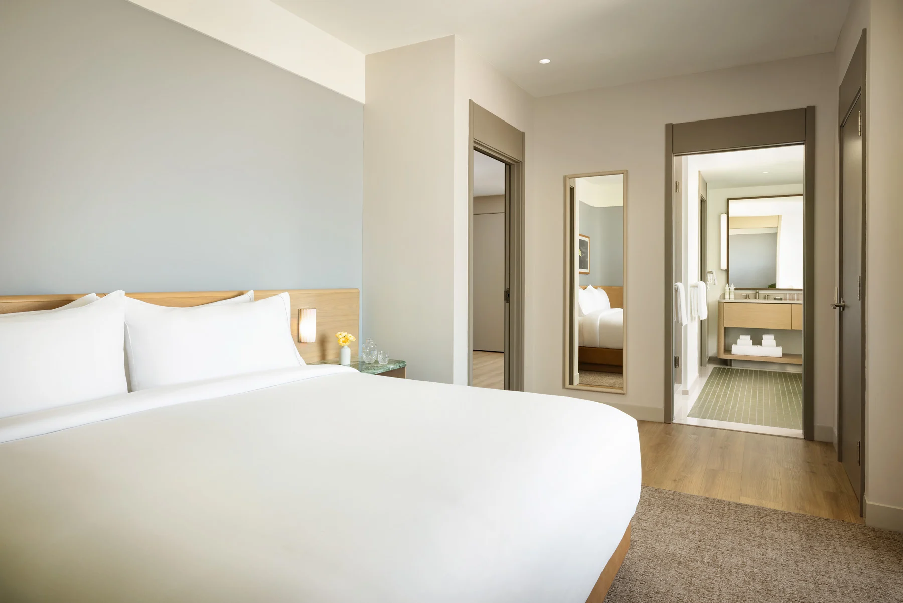 Hotel bedroom with a large white bed, wooden headboard, and a side table with a small vase of yellow flowers; doorway and mirrors on the wall to the right.