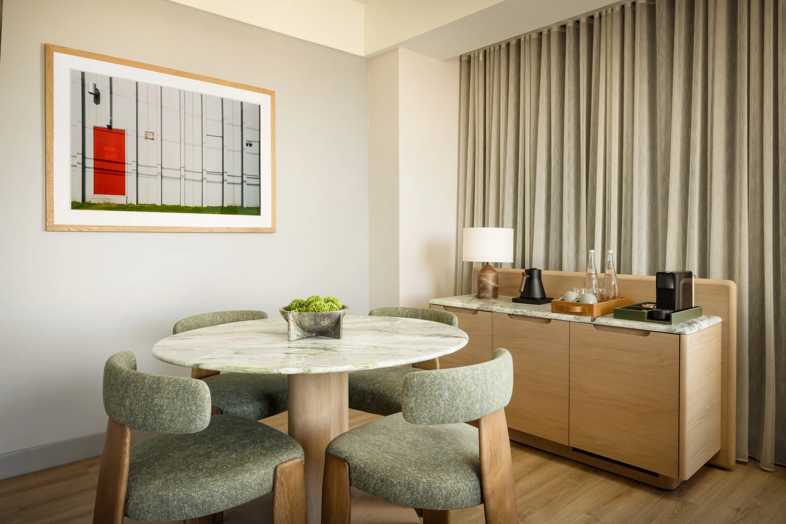 Round marble dining table with green upholstered chairs in a bright, modern room; framed wall art and a curtained wall in the background.