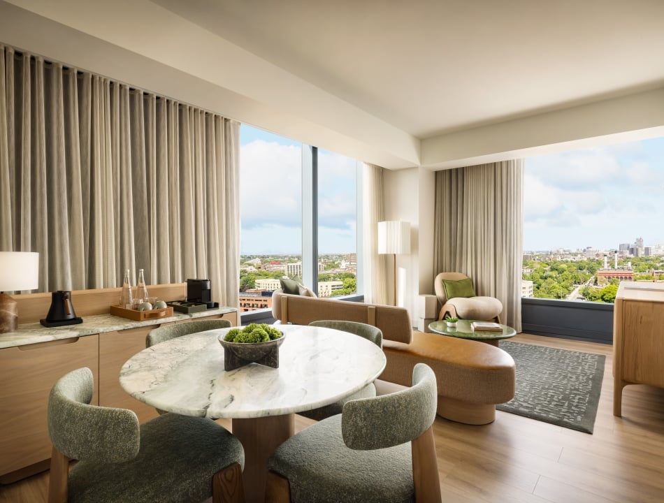 Hotel suite living area with marble round dining table, green upholstered chairs, and a view of the city through large windows.
