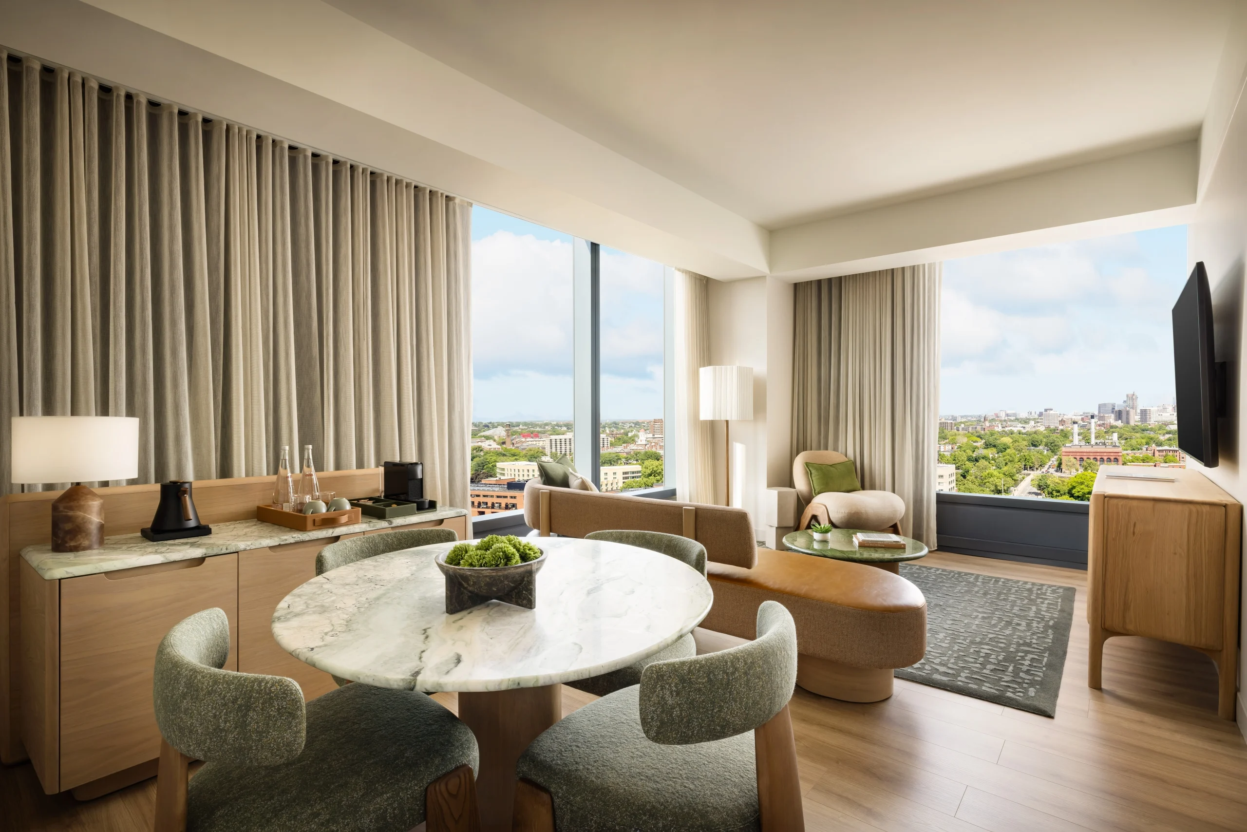 Hotel suite living area with marble round dining table, green upholstered chairs, and a view of the city through large windows.