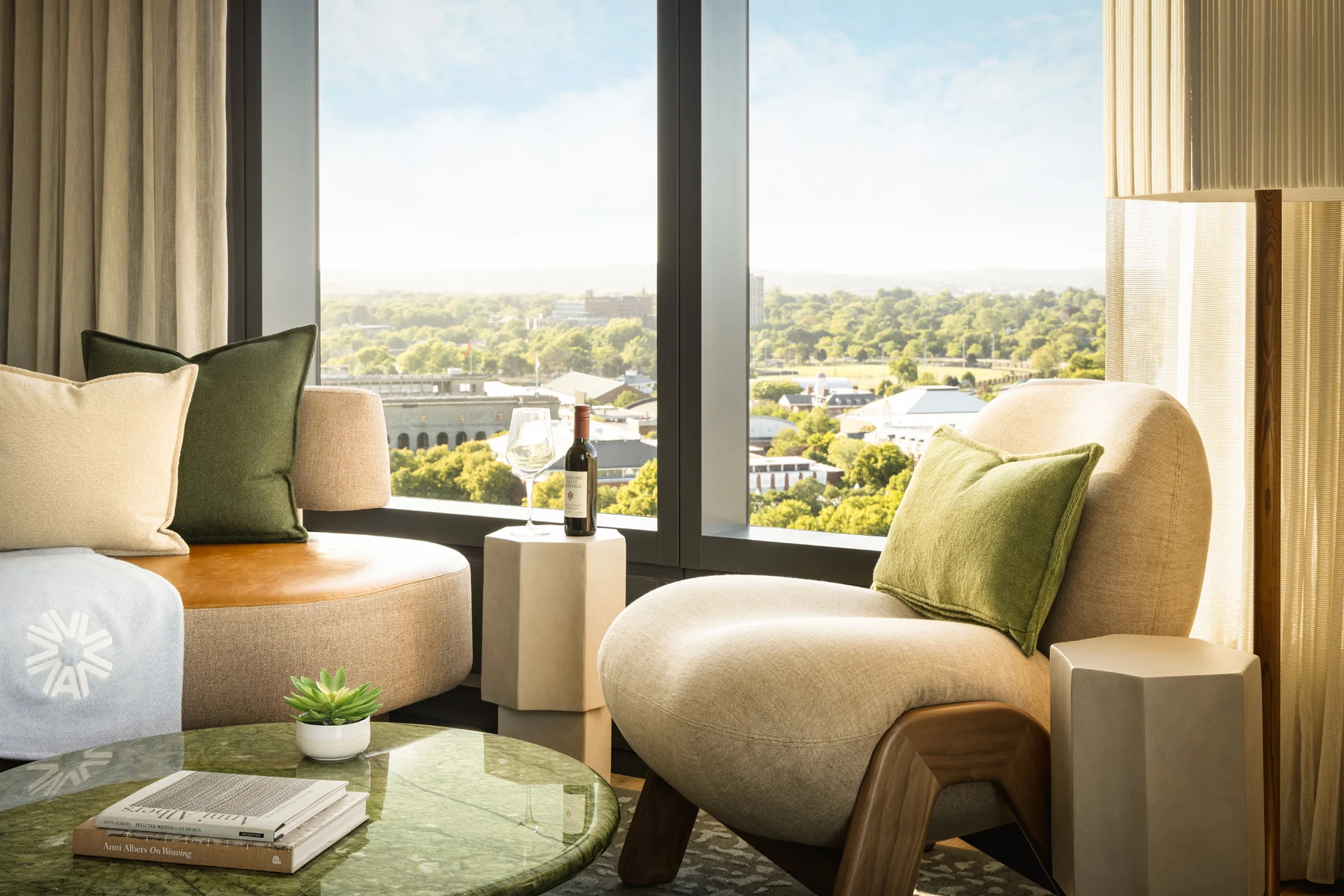 Living room with beige sofa and green cushions by a large window overlooking a cityscape; a bottle of wine and glass on a pedestal beside the sofa.