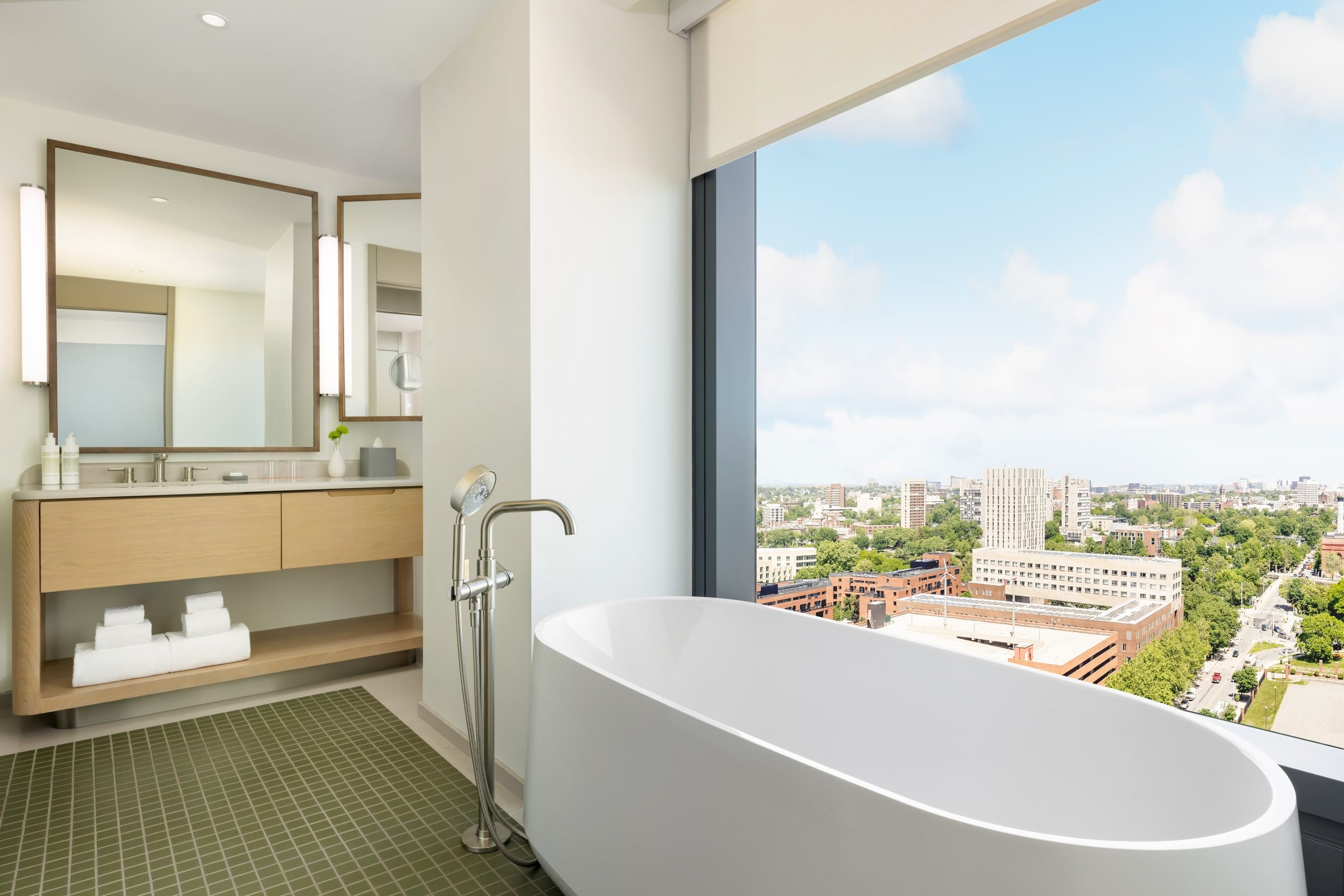 Bright bathroom with a white freestanding tub beside a large window offering a cityscape view; wooden vanity and mirrors reflect soft light.
