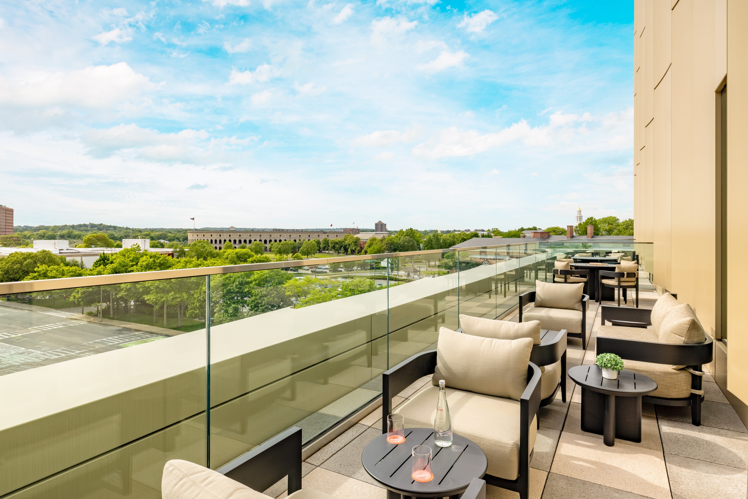 Rooftop balcony lounge with beige cushions, black tables, glass railing, and a cityscape beyond under a blue sky.