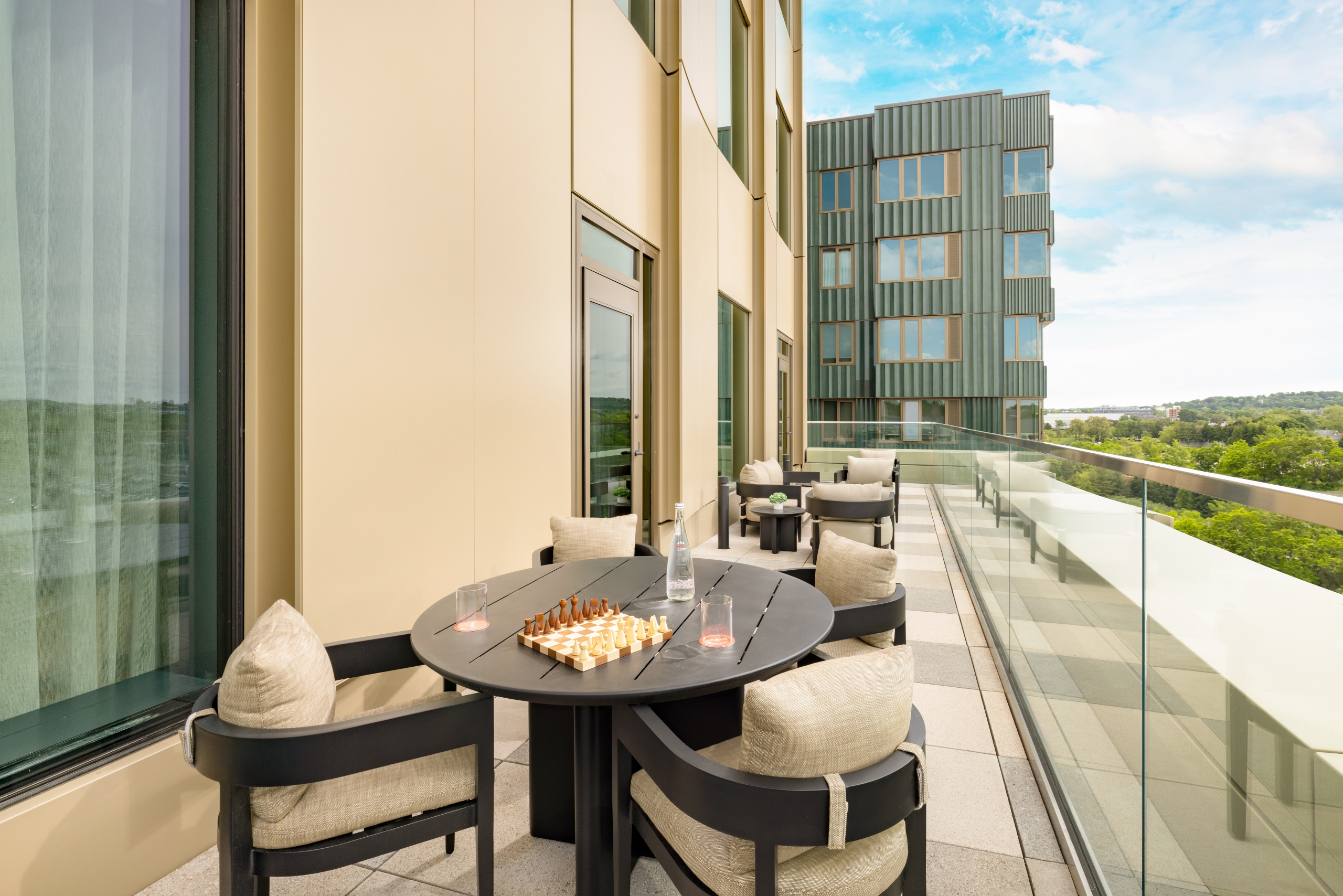 Balcony seating area with a round black table, beige cushioned chairs, a chessboard, and drinks, overlooking a cityscape and greenery.