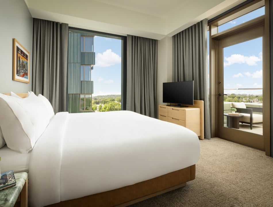 Hotel bedroom with a white bed, grey curtains, wooden dresser with a TV, and a balcony door showing outdoor seating and blue sky.