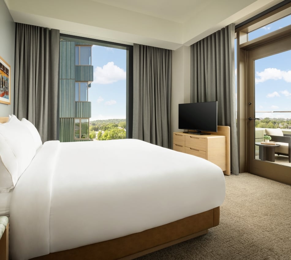 Hotel bedroom with a white bed, grey curtains, wooden dresser with a TV, and a balcony door showing outdoor seating and blue sky.