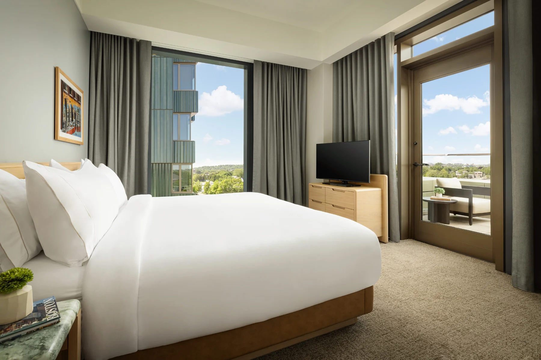 Hotel bedroom with a white bed, grey curtains, wooden dresser with a TV, and a balcony door showing outdoor seating and blue sky.