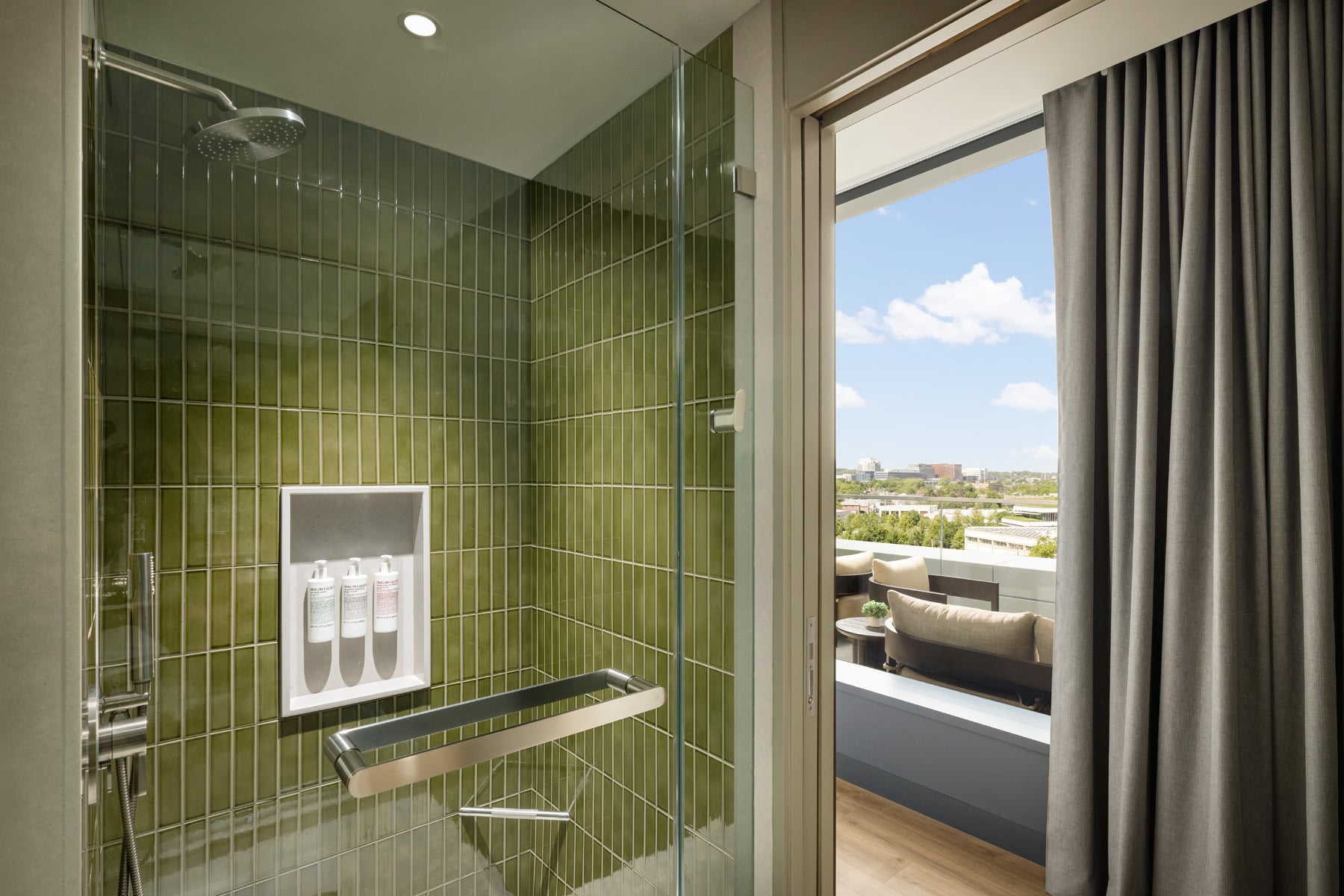 Shower with green tile walls, glass door, and built-in shampoo niche; open door reveals a balcony with seating and a cityscape beyond.