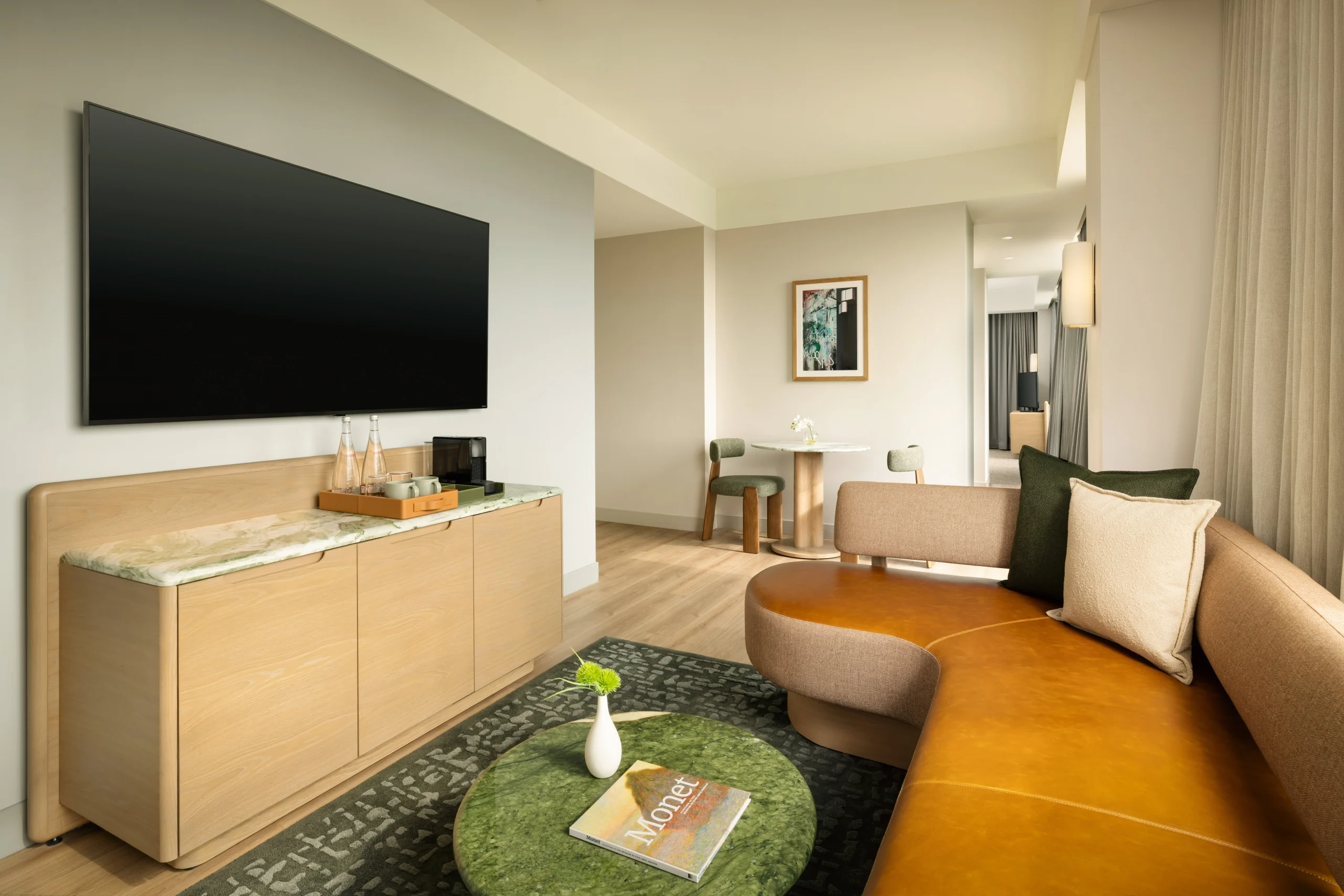Hotel suite living area with a wall-mounted flat-screen TV above a light wood cabinet, green marble coffee tables, and a curved tan leather sofa area carpeted in a dark patterned rug. A Monet magazine rests on one table.