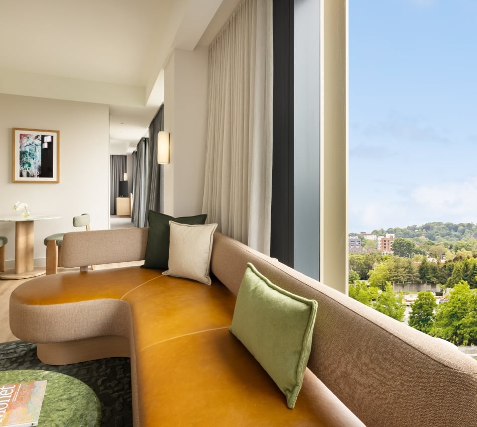 Modern living area with a curved tan sofa, green cushions, and a round leather ottoman, overlooking a city and green treetops through large windows.