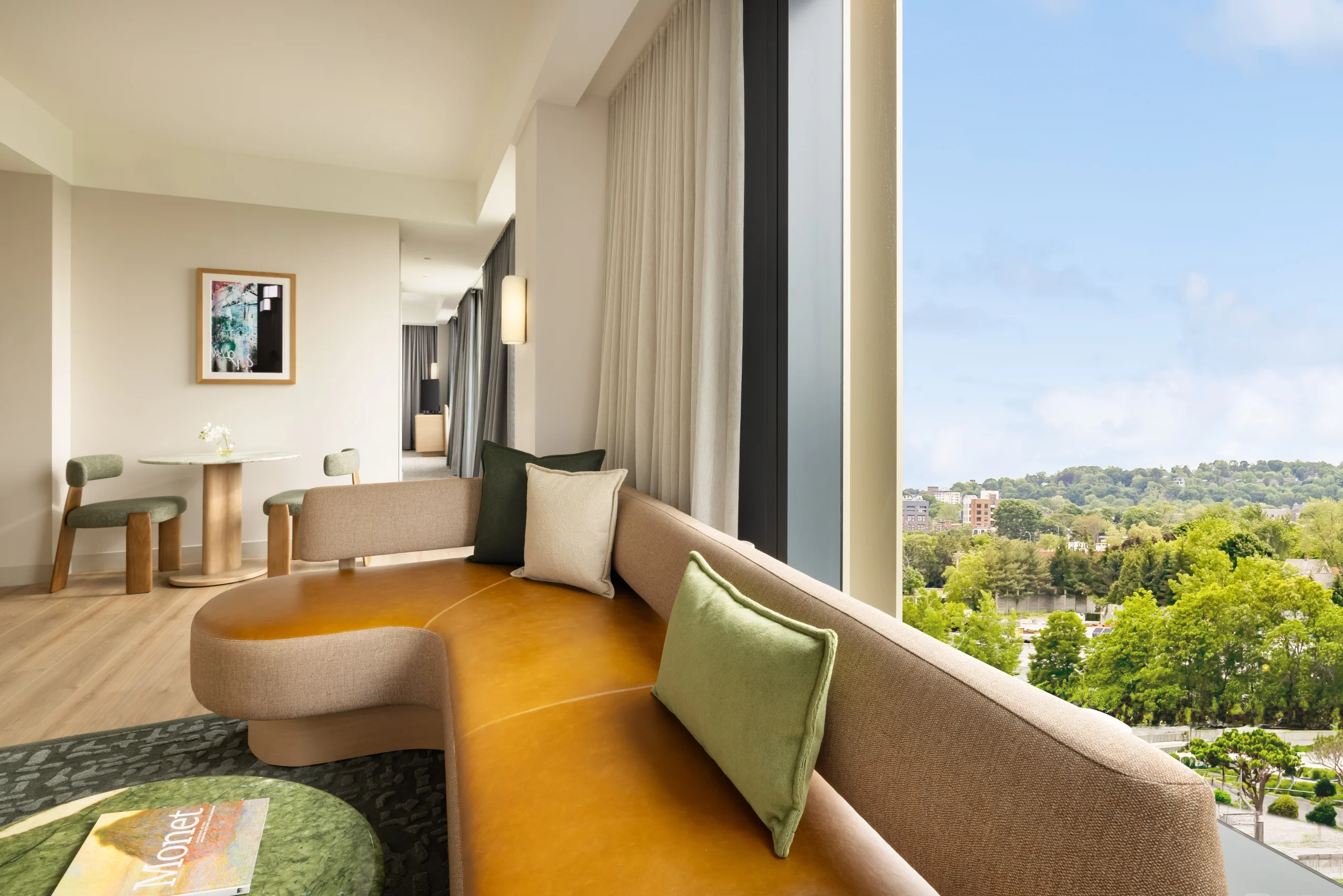 Modern living area with a curved tan sofa, green cushions, and a round leather ottoman, overlooking a city and green treetops through large windows.