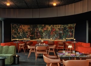 A moody lounge with a curved red banquette, tan chairs, marble tables, and a large figurative tapestry spanning the dark curved wall.
