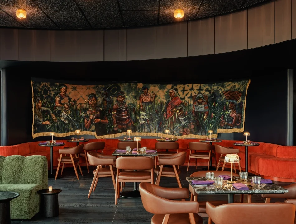 A moody lounge with a curved red banquette, tan chairs, marble tables, and a large figurative tapestry spanning the dark curved wall.