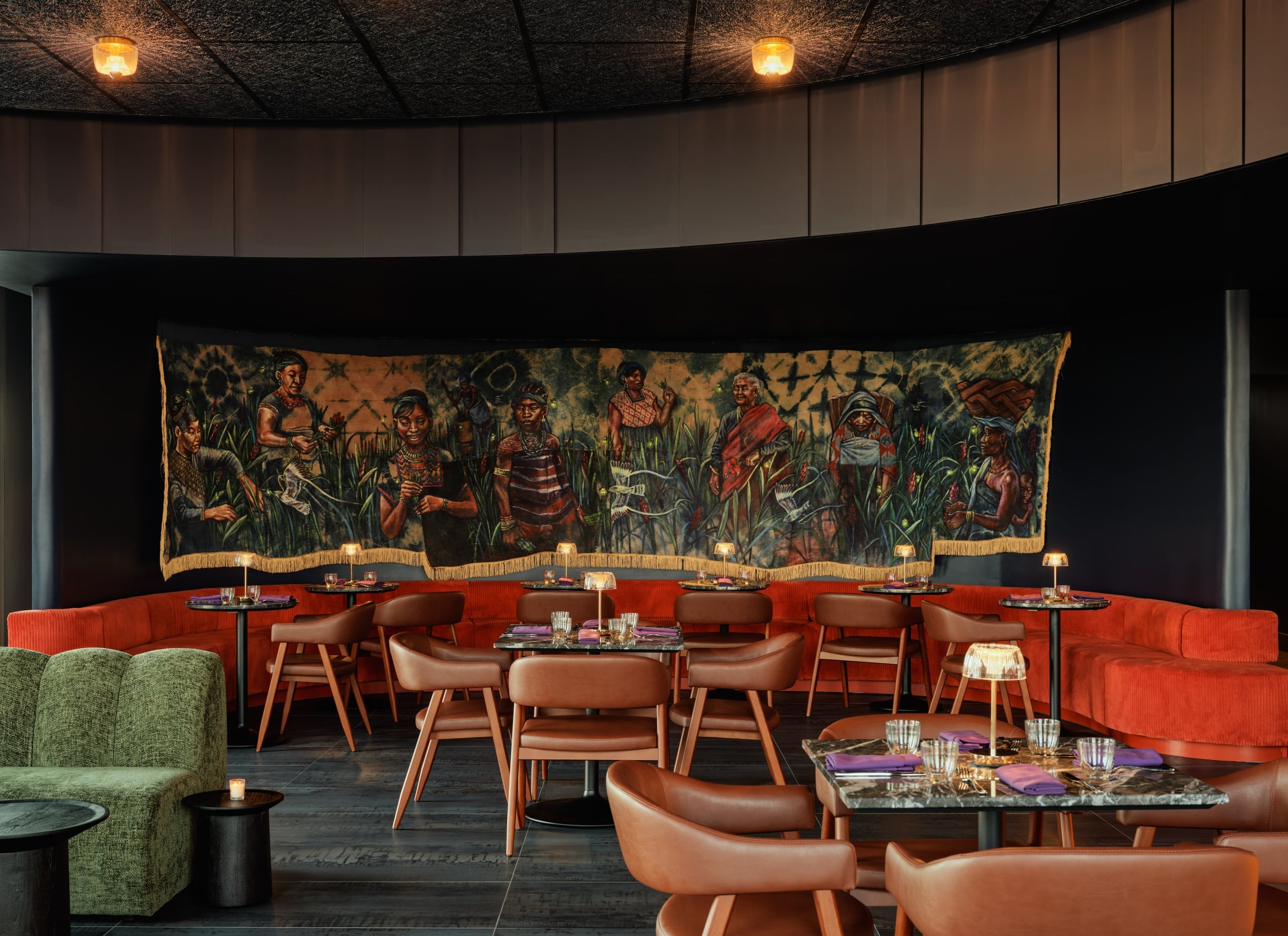 A moody lounge with a curved red banquette, tan chairs, marble tables, and a large figurative tapestry spanning the dark curved wall.