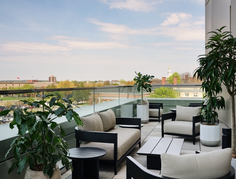 Modern rooftop terrace with black-framed seating, beige cushions, and large potted plants overlooking a city skyline.