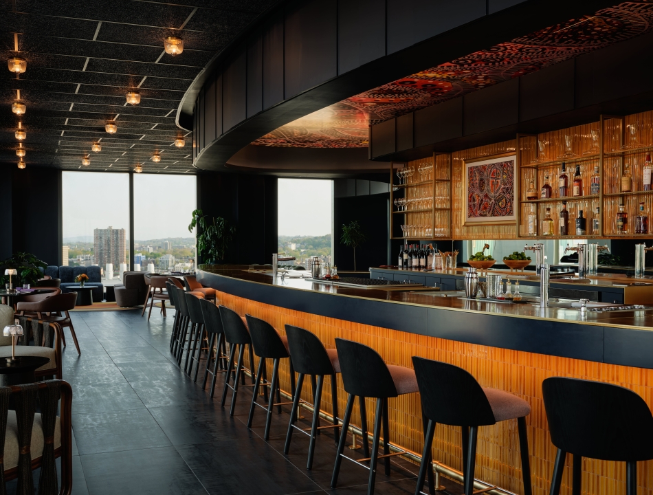 Modern bar interior with a long curved counter, black stools, and warm amber lighting; orange paneling along the bar and glass shelves behind it show bottles and glassware, with a city view through large windows in the background.