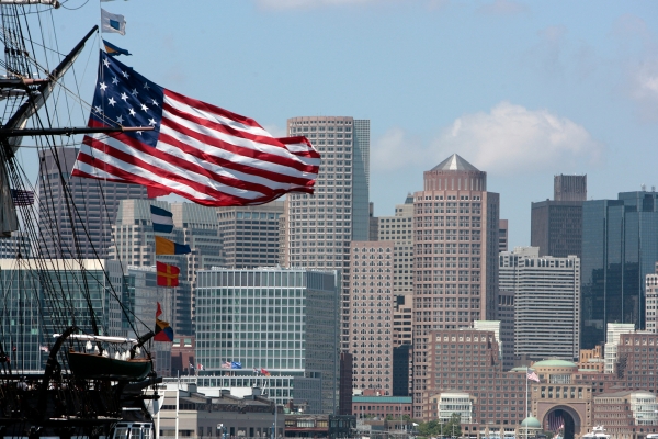 American flag billowing from a tall ship's rigging with a city skyline in the background.