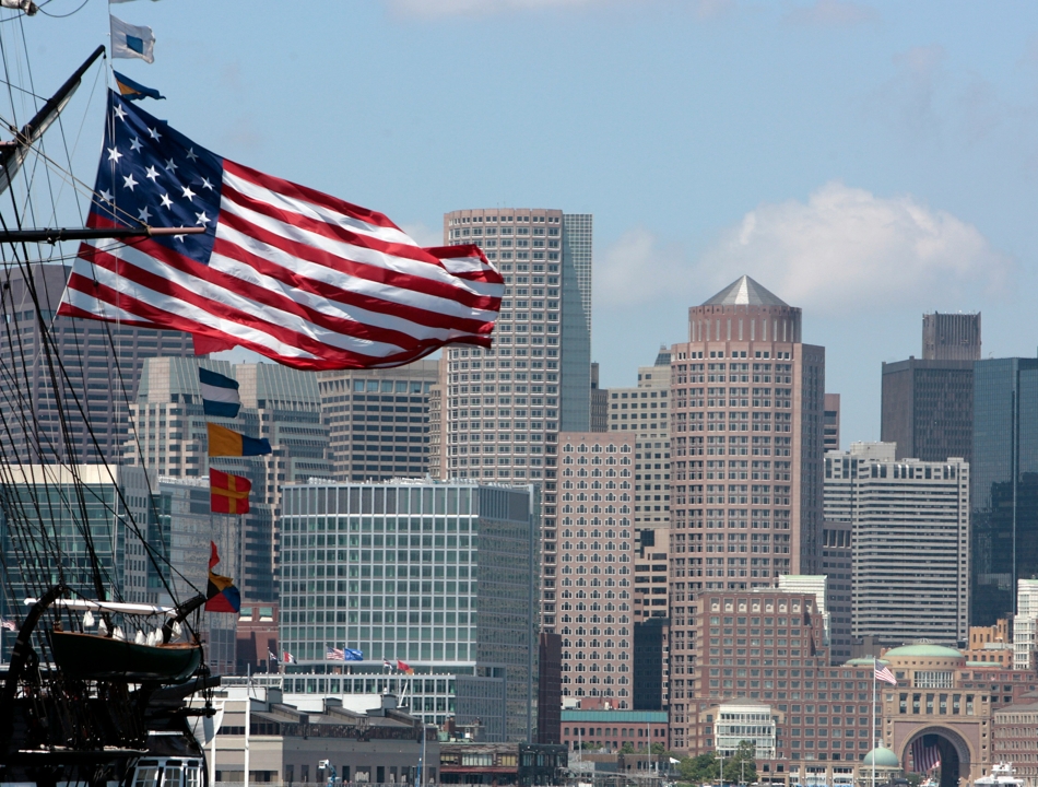 American flag billowing from a tall ship's rigging with a city skyline in the background.