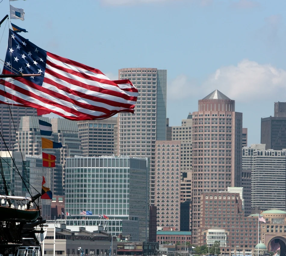 American flag billowing from a tall ship's rigging with a city skyline in the background.