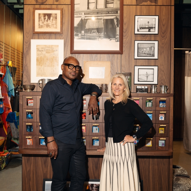 Two people pose in a vintage sports shop, standing in front of a wooden wall with framed photos and shelves of small items behind them.