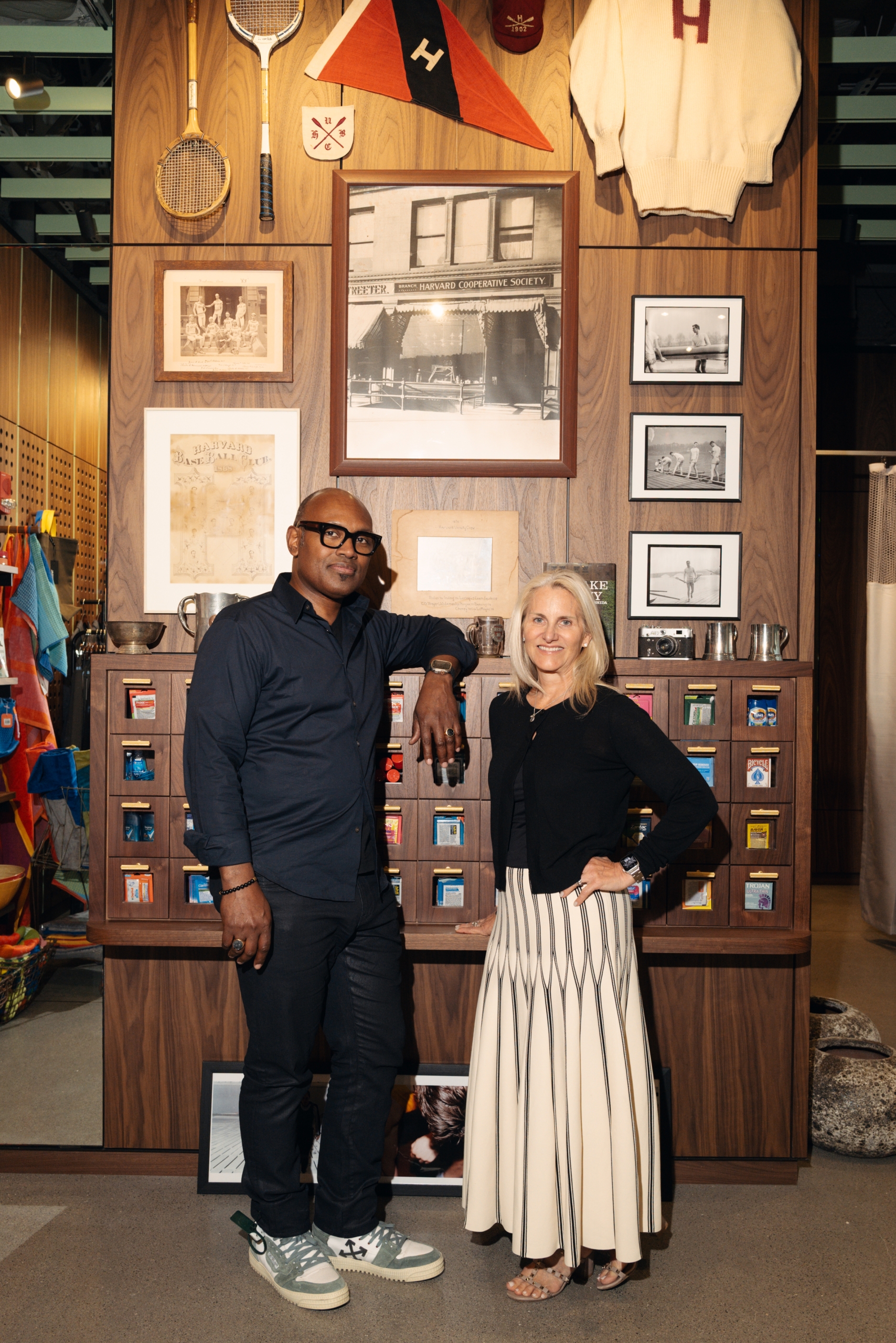 Two people pose in a vintage sports shop, standing in front of a wooden wall with framed photos and shelves of small items behind them.