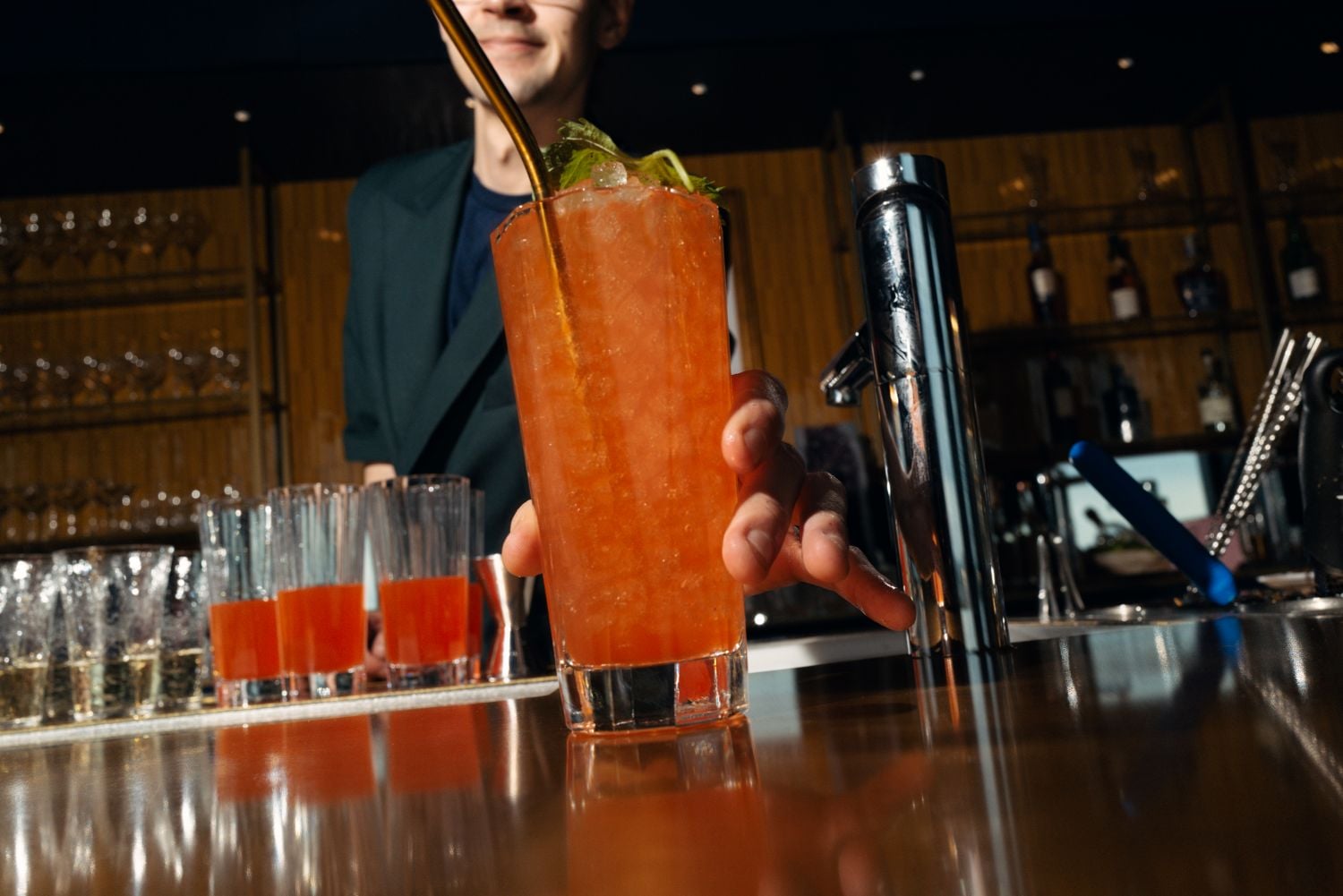 Bartender hands a tall orange cocktail to a customer at a bar, garnished with mint leaves.