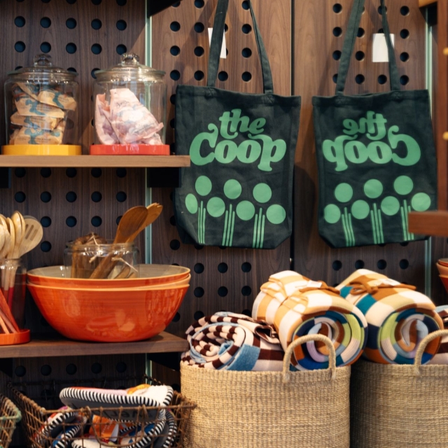 Store display with two black tote bags reading 'the coop' in green, hanging on a wooden pegboard, surrounded by jars, bowls, blankets and woven baskets.