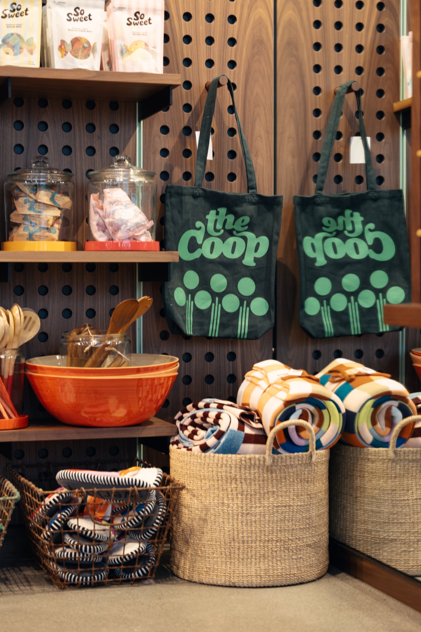 Two dark green tote bags with a teal 'the coop' logo hang on a wooden pegboard display in a gift shop area.