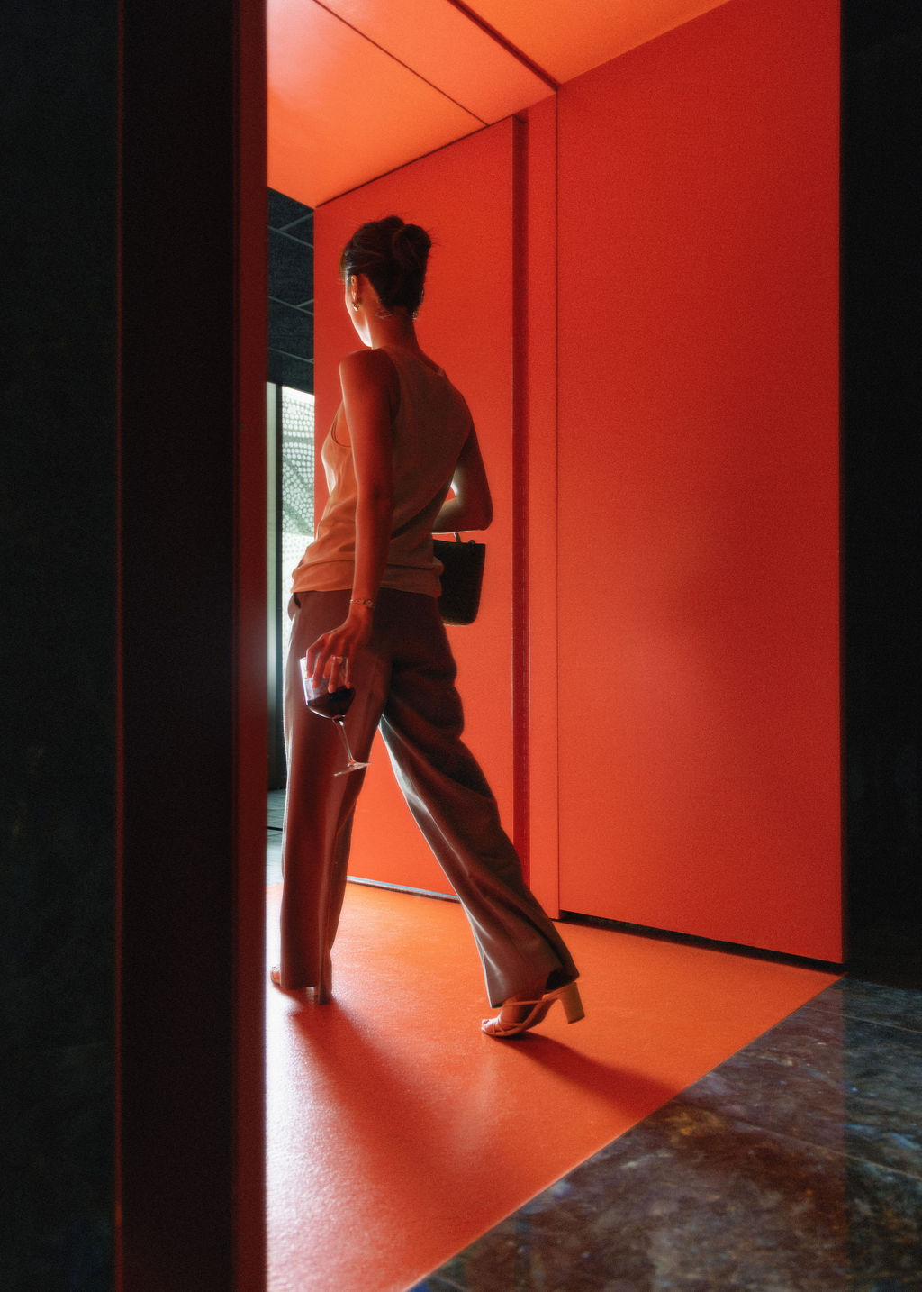 Woman with a sleeveless top and wide-leg pants stands in a red-lit doorway, holding a wine glass.